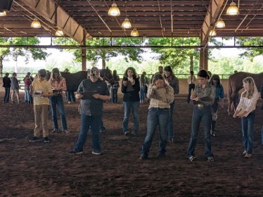 Group of people standing in an indoor arena, looking down at papers while horses stand in the background.