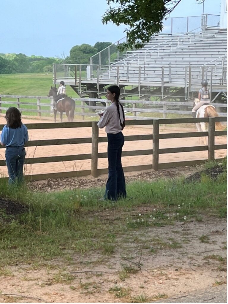 Two people watch riders on horses in a fenced outdoor arena with bleachers behind them.